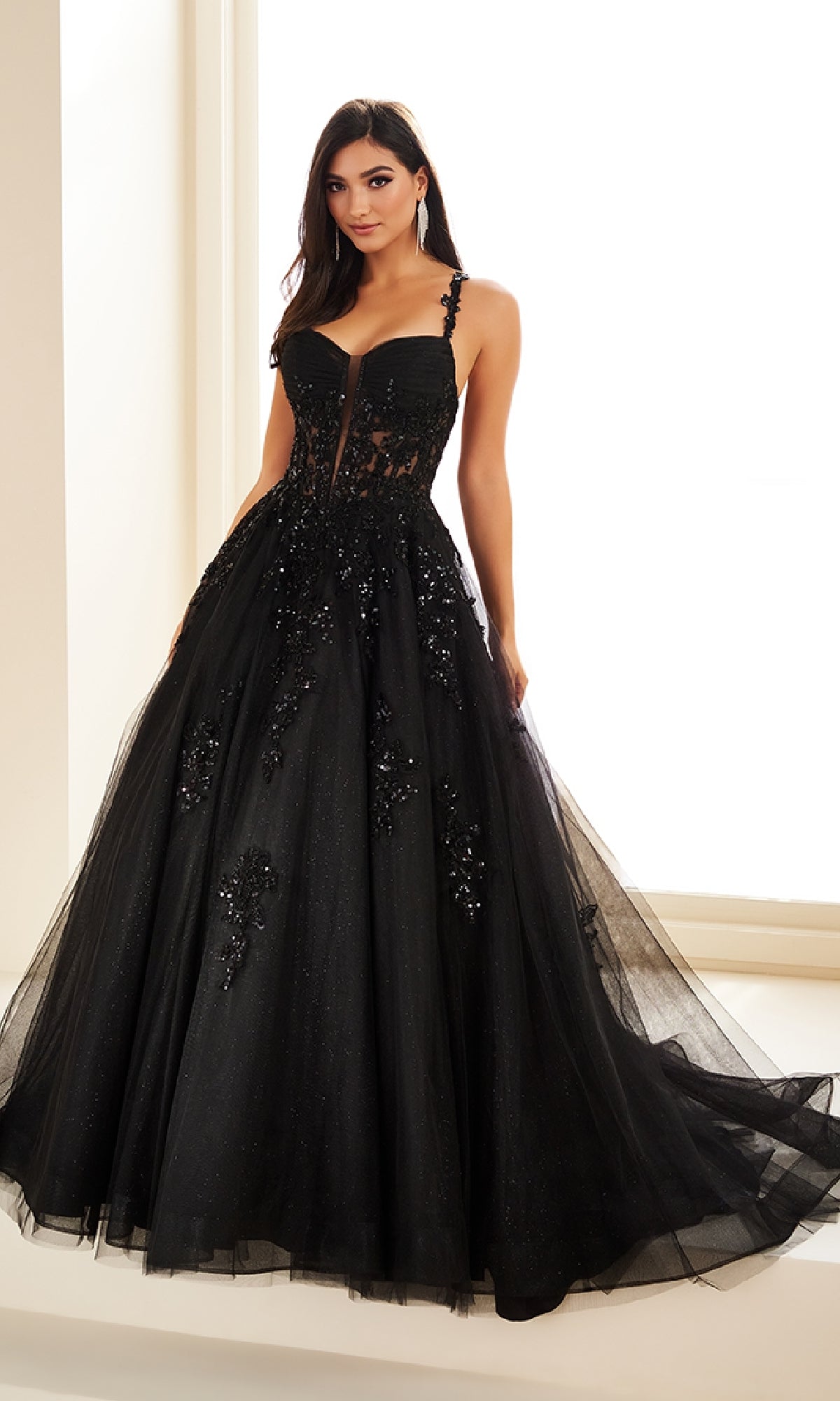A woman stands indoors wearing the Ellie Wilde EW36202 long prom dress, a strapless black ball gown featuring a corset bodice, floral embellishments, and a tulle skirt.